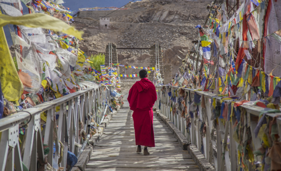 Tibetan monk wearing a red robe walking across a bridge covered with multicolor prayer flags.